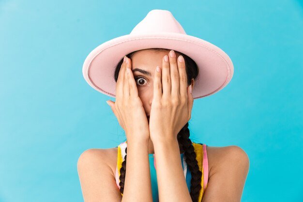 Image closeup of scared caucasian woman wearing hat covering her face and looking at camera isolated over blue wall