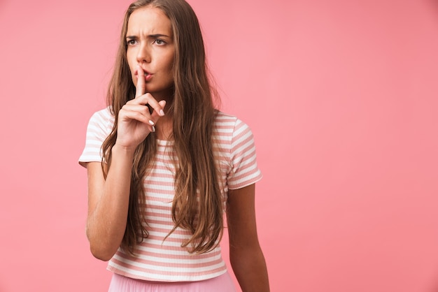 Image closeup of strict young girl frowning and showing hush gesture with keeping finger at lips isolated over pink wall