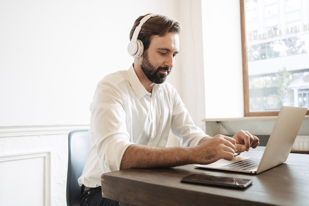 Image of concentrated bearded businessman wearing white shirt using headphones while working at laptop in office