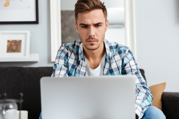 自宅のソファに座ってラップトップコンピューターを使用して、檻のプリントでシャツを着た集中した剛毛の男性の画像。