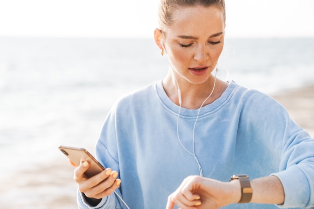 Image of a concentrated fitness woman outdoors on beach using mobile phone listening music with earphones looking at watch clock.