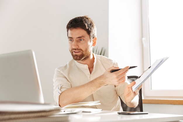 Image of concentrated office guy 30s wearing white shirt using laptop and paper documents, while working at modern workplace