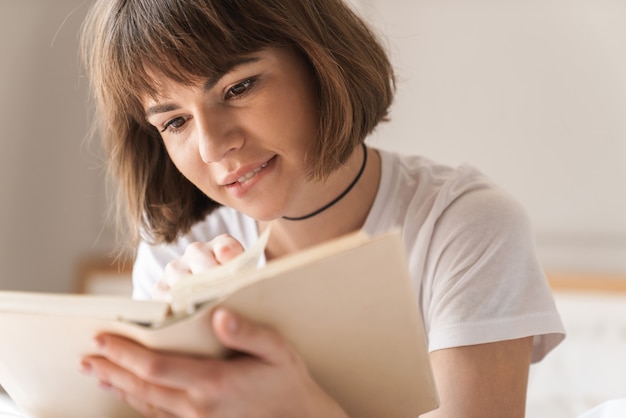 自宅の屋内で集中してリラックスした若い美しい女性の画像は、本を読んでベッドに横たわっています。