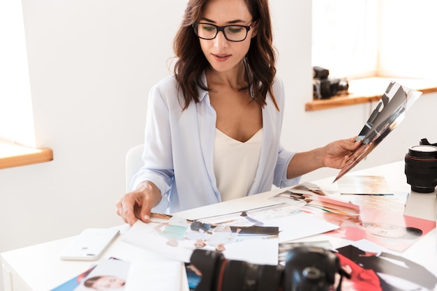 Image of a concentrated young photographer woman in office sitting at the table holding photos.