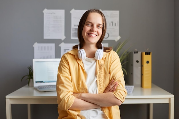 Image of confident hipster woman office worker or student sitting on workplace wearing yellow shirt and headphones over her neck keeps arms folded looking at camera with toothy smile