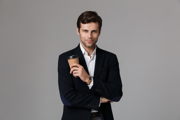 Image of confident man 30s in businesslike suit smiling and holding coffee, isolated over gray wall