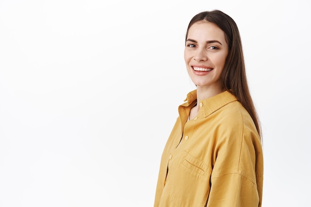 Image of confident young woman with professional and determined face, standing half turned, turn head at camera and smile happy, standing over white background