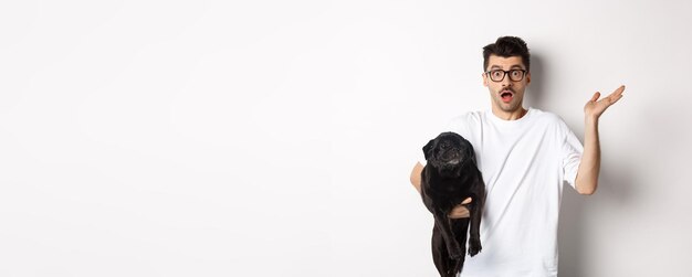 Изображение растерянного хипстера, держащего собаку и пожимающего плечами, озадаченно поднимающего руку, стоящего с