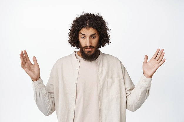 Image of confused middleeaster guy looking down with raised empty hands dropped something below standing in casual outfit over white background