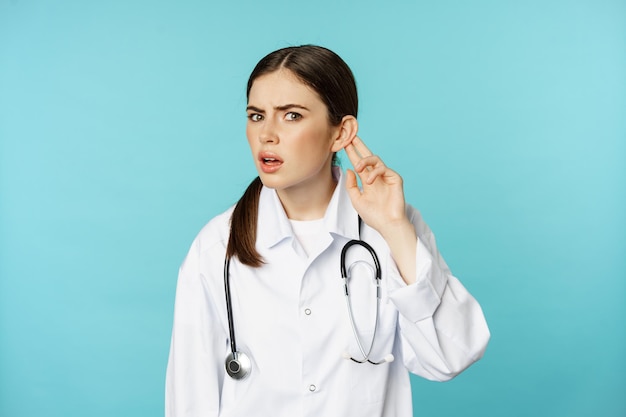Image of confused woman doctor cant hear you, holding hand near ear and looking puzzled, speak louder gesture, torquoise background.