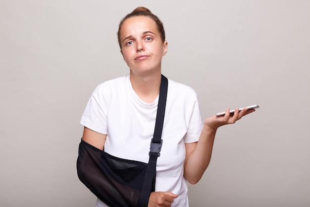 Image of confused woman wearing casual Tshirt standing in arm sling holding cell phone doesn't know where is hospital finding information in the internet posing isolated on light gray background