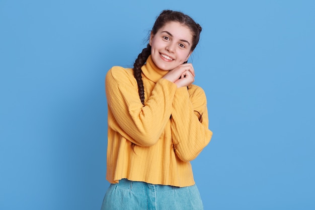 Image of cute european girl with two braids wearing yellow sweater, looks smiling, looking at front