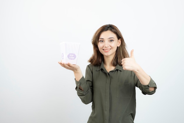 Immagine di una ragazza carina con una scatola di popcorn che posa alla macchina fotografica e dà i pollici in su. foto di alta qualità