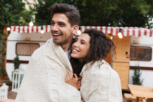 Изображение милой счастливой положительной любящей пары мужчины и женщины, обнимающей возле передвижного дома трейлера в пледе.