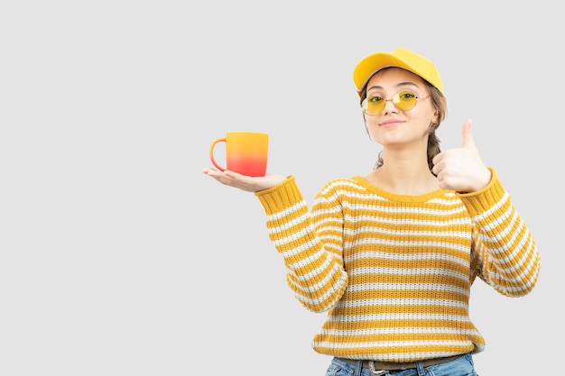 Image of cute young woman in glasses showing thumb up and holding a mug . High quality photo