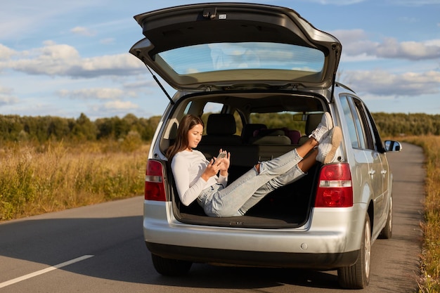 Image of dark haired Caucasian young adult woman sitting in car trunk female wearing white shirt and jeans resting while traveling and using cell phone enjoying good weather and her journey