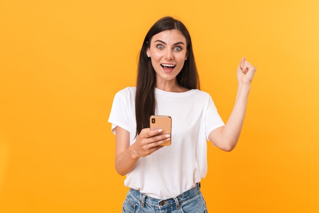 Image of delighted brunette woman wearing basic clothes rejoicing and holding cellphone isolated over yellow wall