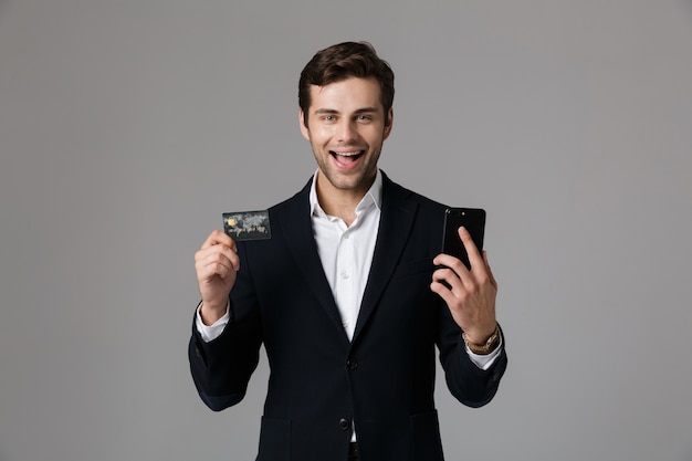 Immagine di felice uomo 30s in tailleur tenendo il telefono cellulare nero e la carta di credito, isolato sopra il muro grigio