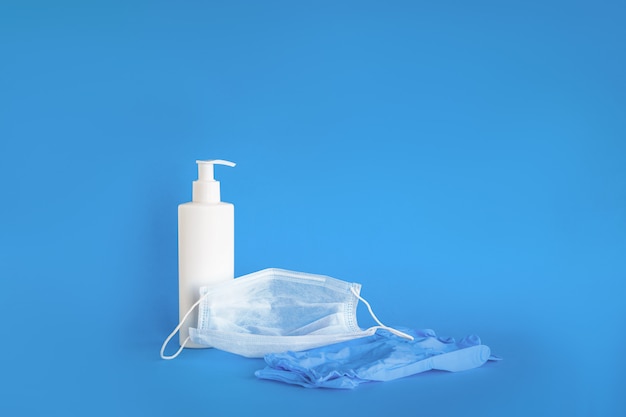 Image of disposable protective medical mask, latex gloves and bottle with sanitizer close-up on a blue wall. Space for text. The concept of protection against the virus, medicine and health