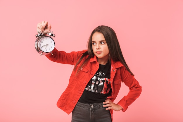 Изображение недовольной девушки 8-9 лет в повседневной одежде, держащей будильник, стоя, изолированной над красной стеной