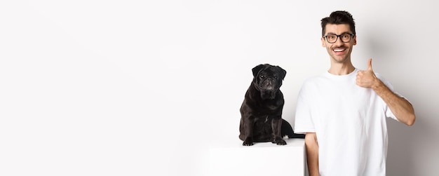 Image of dog owner and cute black pug looking at camera man showing thumbup in approval recommending