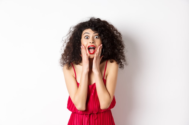 Image of elegant woman scream surprised, staring at camera, see promo offer and shouting of joy, standing in dress over white background.