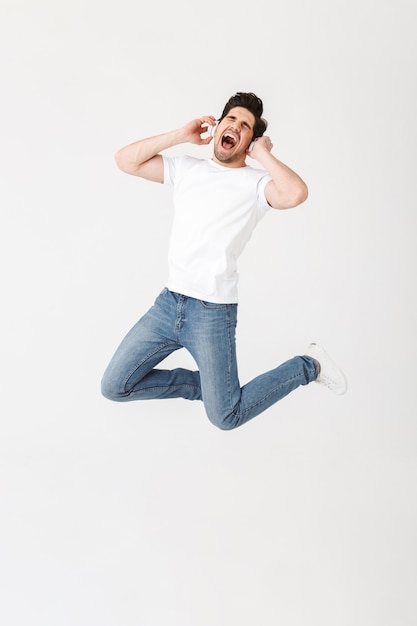 Image of emotional excited young man posing isolated over white wall  listening music with headphones jumping.