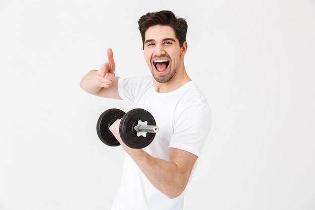 Image of emotional happy excited young man posing isolated over white wall holding dumbbell make exercise pointing.