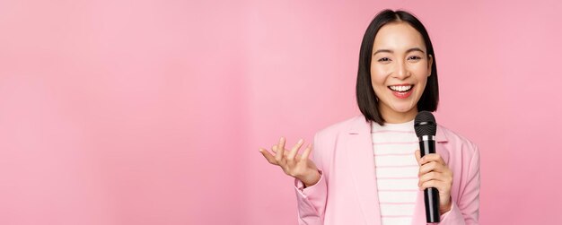 Image of enthusiastic asian businesswoman giving speech talking with microphone holding mic standing in suit against pink studio background person