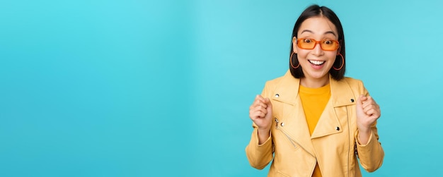 Image of enthusiastic young asian woman celebrating triumphing looking surprised and happy clapping hands satisfied standing over blue background