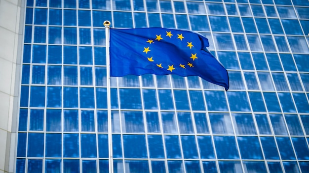 Image of EU flag fluttering on wind against high business office building made of concerete and glass. Concept of ecenomics, development, government and politics