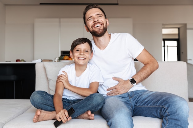 Photo image of european father and son laughing, while sitting on couch indoor with remote control