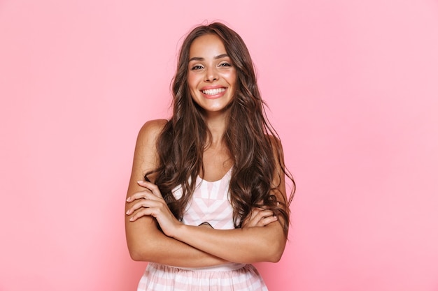 Photo image of european lovely woman 20s with long hair wearing dress smiling at you with arms crossed, isolated over pink wall