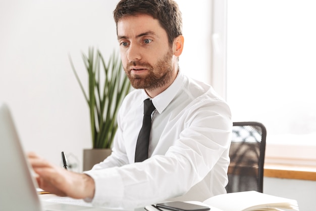 Image of european office worker 30s wearing white shirt working with laptop and notebook, in modern workplace