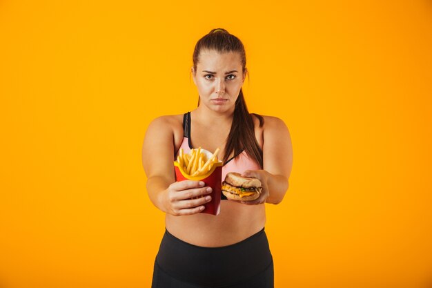 Foto immagine della donna sovrappeso europea in tuta che fa gesto di arresto mentre si tiene panino e patatine fritte, isolato su sfondo giallo