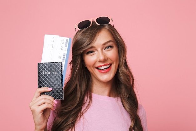 Image of european woman having beautiful brown locks smiling while holding passport and air tickets, isolated over pink background