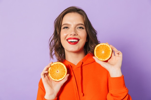 Image of an excited beautiful young woman posing isolated over purple wall wall holding orange citrus