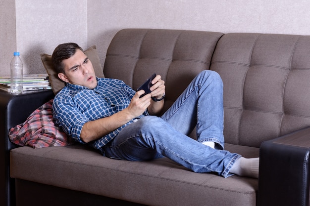 Image Excited of a bristle man dressed in shirt in a cage print, jeans and white socks, lies on sofa in home and playing at smatphone. Passionate about the game