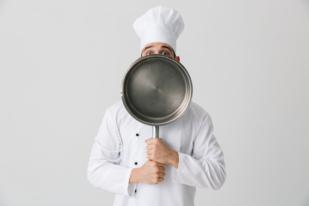 Image of excited emotional young man chef indoors isolated over white wall background holding frying pan.