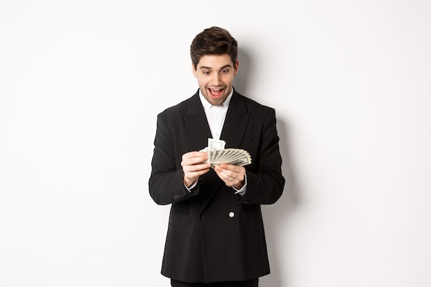 Image of excited handsome businessman, counting money and smiling amused, standing against white background in suit.