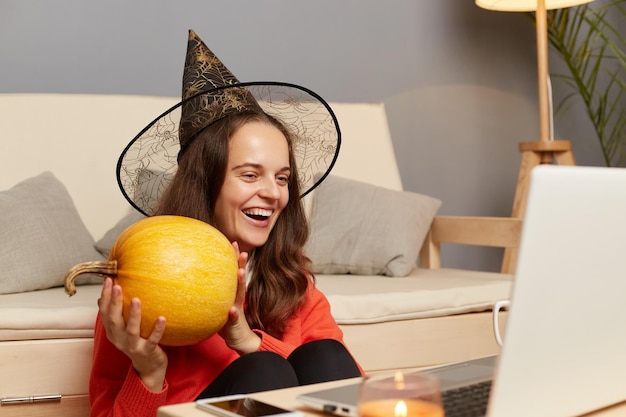 Image of excited happy optimistic young adult woman in witch hat having online video call on laptop celebrating halloween at home showing pumpkin to notebook display