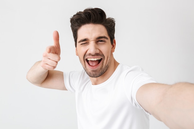 L'immagine di un giovane uomo felice eccitato in posa isolato su un muro bianco fa un selfie con la telecamera che punta a te.