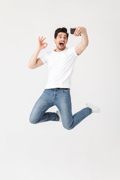 Image of excited happy young man posing isolated over white wall  using mobile phone take a selfie showing okay gesture.