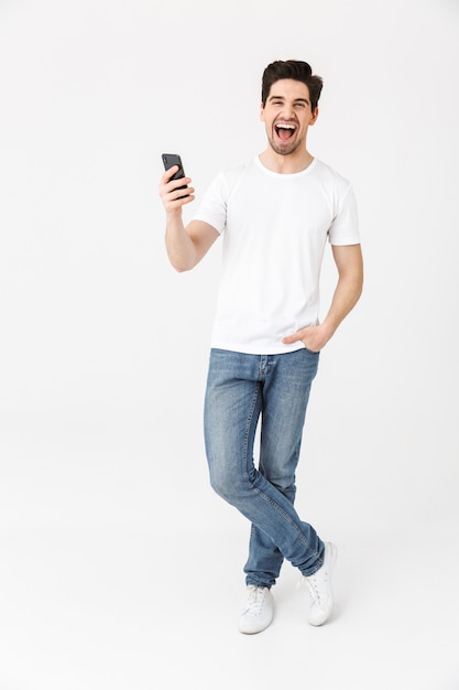 Image of excited happy young man posing isolated over white wall  using mobile phone.