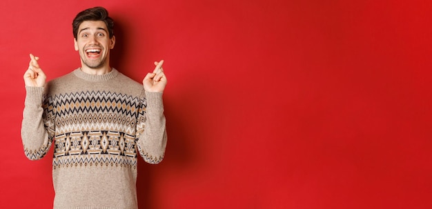 Image of excited and hopeful young man making wish wearing winter sweater crossing fingers for good luck and smiling anticipating christmas eve standing over red background