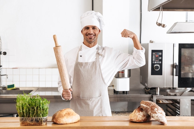 Image of excited man baker in white uniform smiling, while standing at bakery with bread on table