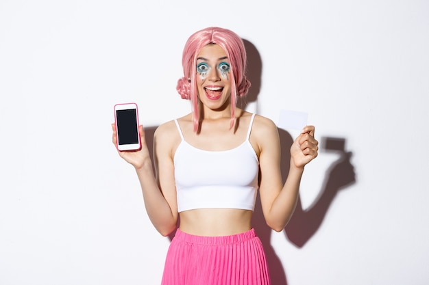 Image of excited party girl in pink wig and halloween outfit showing credit card and smartphone scre...