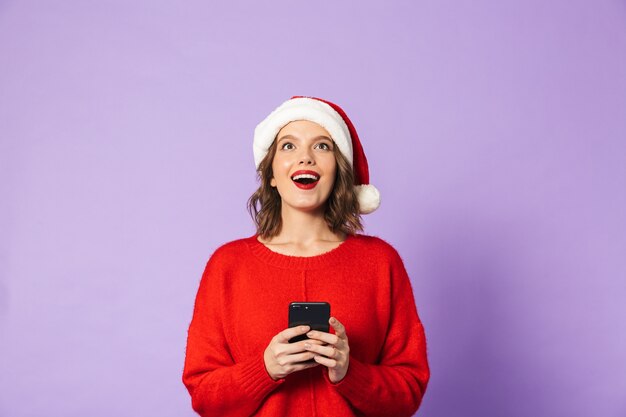 휴대 전화를 사용 하여 보라색 벽 위에 절연 크리스마스 모자를 쓰고 흥분된 충격 된 젊은 여자의 이미지.