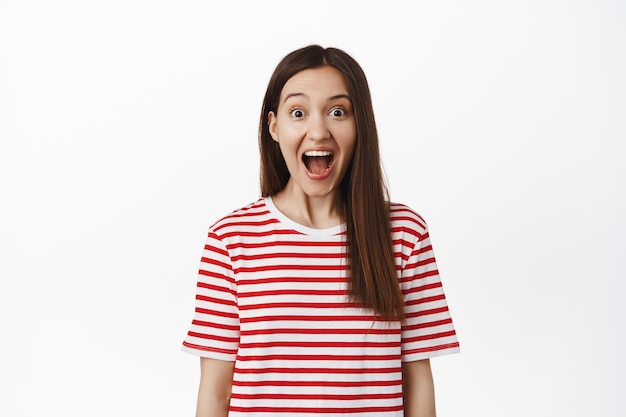 Image of excited surprised girl drop jaw, scream from amazement and looking at camera amazed, staring in awe at surprise for her, standing in striped t-shirt against white background.