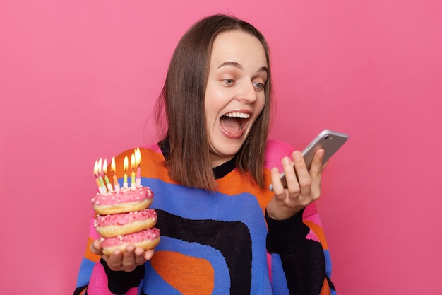 興奮した女性のイメージは、ろうそくを燃やしておいしいドーナツを保持し、バラの背景に孤立して立っている誕生日を祝うセーターを着てお祝いのメッセージをスマートフォンで読んでいます